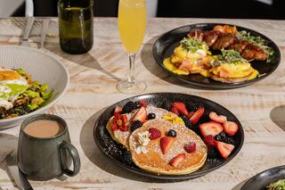 A spread featuring fluffy pancakes topped with berries and flowers, a savory egg dish, a coffee mug, and a mimosa. Bright and inviting.