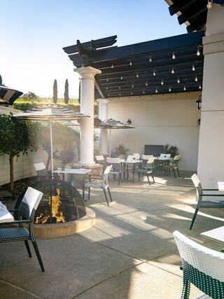 A stylish outdoor dining area features elegant tables, shaded by umbrellas, with a cozy fire pit and lush greenery under a bright sky.