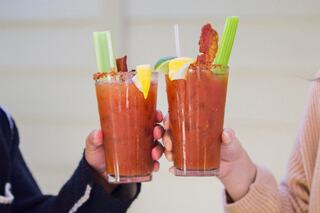 Two hands raise tall glasses of Bloody Marys, garnished with celery, lemon, and bacon, celebrating a festive moment.