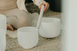 A person in soft, neutral clothing plays crystal singing bowls, creating soothing sounds in a serene, natural setting.