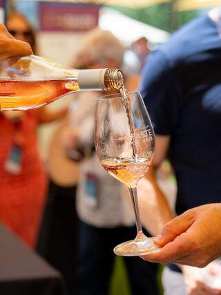 A hand pours rosé wine from a bottle into a glass at a lively outdoor event, with blurred attendees in the background.