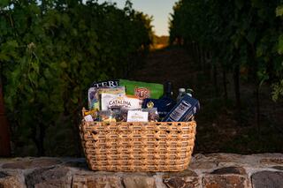 A woven basket filled with gourmet snacks and drinks sits atop a stone wall, surrounded by lush vineyard rows at sunset.