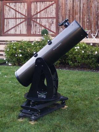 A sleek telescope stands on a lawn, facing upward, with a rustic barn door and flowers in the background, ready for stargazing.