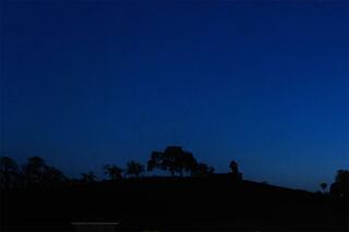 A dark silhouette of trees and a distant figure stands against a deep blue twilight sky, evoking a serene and tranquil atmosphere.