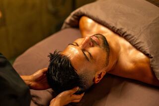 A man relaxes on a massage table, receiving a soothing head massage in a tranquil setting, showcasing tranquility and self-care.