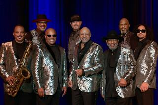 A group of eight musicians poses together, all dressed in shiny silver jackets, showcasing a blend of instruments and styles on stage.