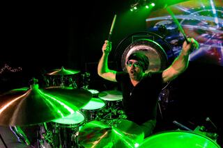 A drummer passionately performs, playing with intensity under vibrant green stage lights, surrounded by a drum set and a large gong.