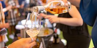 A hand pours rosé wine into a glass at a lively event, with blurred figures in the background enjoying the atmosphere.