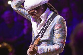 A musician with long dreadlocks wears a stylish floral suit and a white hat, passionately playing guitar on stage.