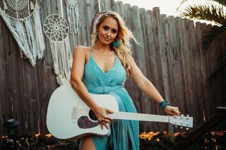 A woman in a flowing turquoise dress holds a white guitar, standing against a wooden fence adorned with dreamcatchers.