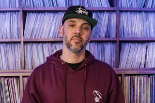 A man stands in front of shelves filled with vinyl records, wearing a maroon hoodie and a black cap, exuding a cool, laid-back vibe.