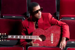 A stylish musician in a red suit and sunglasses poses with a sparkling red guitar amidst velvet theater seats.