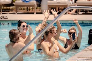 A group of friends joyfully celebrates in a pool, raising their hands and laughing, enjoying a sunny day together.