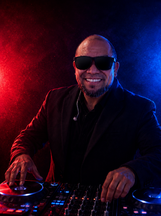 A DJ in sunglasses smiles while mixing tracks, surrounded by vibrant red and blue lights, exuding energy and confidence.