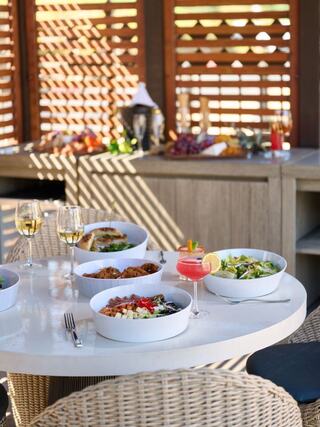 A stylish dining setup features bowls of colorful dishes, cocktails, and glasses of white wine, illuminated by warm sunlight.