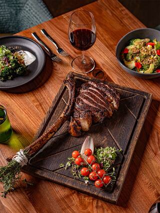 A beautifully arranged feast featuring a grilled tomahawk steak, vibrant cherry tomatoes, and fresh herbs, complemented by a glass of red wine.