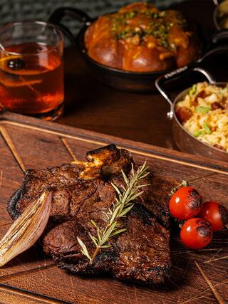 Juicy grilled steak garnished with rosemary and cherry tomatoes, accompanied by a refreshing drink and savory side dishes on a wooden board.