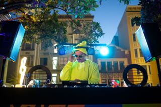 A DJ in a bright yellow outfit plays music outdoors at dusk, surrounded by vibrant lights and a lively urban backdrop.
