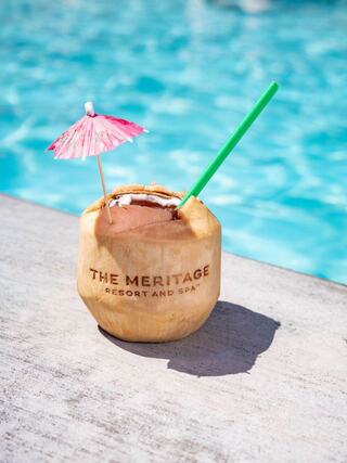 A coconut drink garnished with a pink umbrella and a green straw sits on a poolside ledge, radiating a tropical vibe.