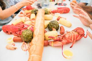A vibrant seafood feast with lobsters, shrimp, artichokes, corn, and a large loaf of bread, enjoyed by hands reaching in for bites.