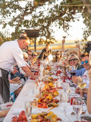 A festive outdoor gathering with a long table set for a seafood feast, as guests enjoy food and drinks under string lights.
