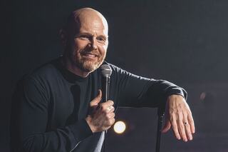A smiling comedian stands on stage, microphone in hand, engaging the audience with a relaxed demeanor. Soft lighting creates an intimate atmosphere.