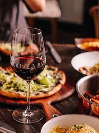 A glass of red wine sits beside a pizza topped with greens, alongside bowls of various dishes, creating a cozy dining setting.