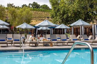A couple relaxes poolside, enjoying drinks and snacks under umbrellas, with a vineyard backdrop and lounge chairs nearby.
