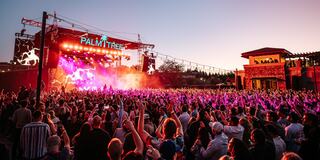 A lively crowd enjoys a concert at sunset, with colorful lights illuminating the stage and vibrant energy in the atmosphere.