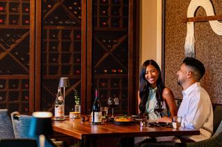 A couple enjoys a cozy dinner with wine, smiling and chatting in an elegant setting with a wine rack in the background.