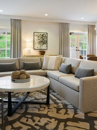 A cozy living room features a sectional sofa, circular coffee table, decorative cushions, and large windows with soft curtains.