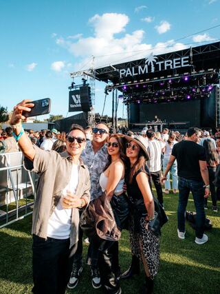 A group of four friends poses for a selfie at a lively outdoor music festival, with a stage and crowd in the background under a blue sky.