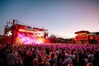 A vibrant crowd enjoys a live performance at dusk, with colorful lights illuminating the stage and surrounding palm trees.