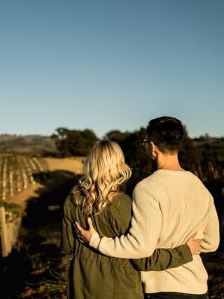 A couple stands close together, overlooking a vineyard under a clear blue sky, sharing an intimate moment amidst nature.