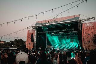 A vibrant outdoor concert scene with a large crowd enjoying a performance while colorful lights illuminate the stage at sunset.