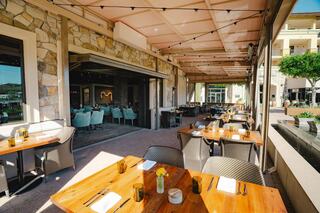 A sunny patio features wooden tables set for dining, with stylish chairs and a backdrop of a modern stone building and greenery.