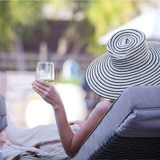 A person in a wide-brimmed, striped hat relaxes on a lounge chair, holding a glass of white wine in a serene outdoor setting.