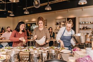 Three women prepare elegant dishes in a modern kitchen, while a group observes eagerly in the background, highlighting a culinary workshop.