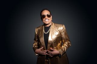 A stylish individual in a gold suit and sunglasses smiles confidently against a dark background, exuding charisma and flair.