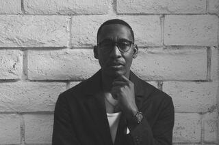 A thoughtful man in glasses poses against a textured white brick wall, wearing a dark jacket and displaying a contemplative expression.