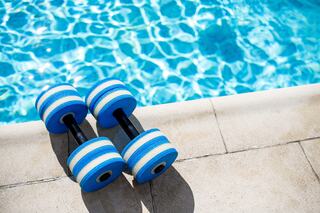 A pair of blue and black striped dumbbells rests on a stone edge beside a sparkling swimming pool.