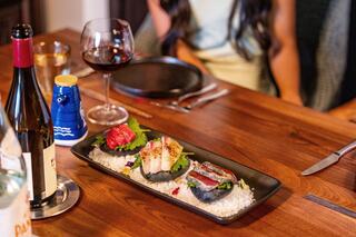 A gourmet dish featuring sushi on a black plate, garnished with greenery and served alongside wine and condiments on a wooden table.