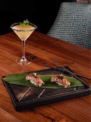 A sleek wooden platter holds two pieces of sushi garnished with gold flakes, accompanied by a stylish cocktail with a salted rim.
