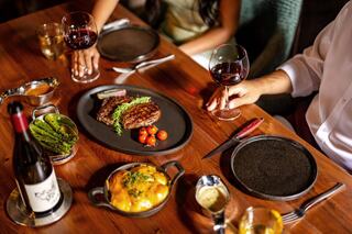 A lavish dinner spread featuring steaks, grilled vegetables, and a creamy dish, accompanied by glasses of red wine on a wooden table.