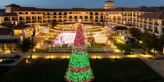 A festive outdoor scene features a large, decorated Christmas tree, string lights, and a hotel backdrop, creating a vibrant holiday atmosphere.