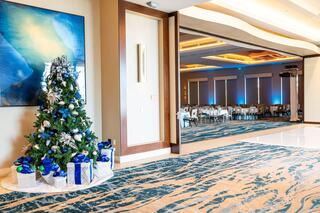 A decorated Christmas tree stands in a bright, spacious room, with festive blue and silver accents, leading to a banquet area.