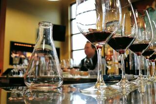A laboratory flask sits on a bar, surrounded by elegant wine glasses filled with red wine, with a blurred figure in the background.