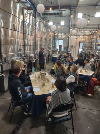 A group of people sits at tables in a spacious venue, listening to a speaker. Stainless steel tanks and decorative lights create an inviting atmosphere.