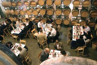 A group of people dining at long tables in a rustic setting filled with wine barrels, engaged in conversation and enjoying their meal.