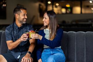 A happy couple shares a toast with colorful drinks, smiling joyfully while seated on a cozy couch in a warm, inviting setting.